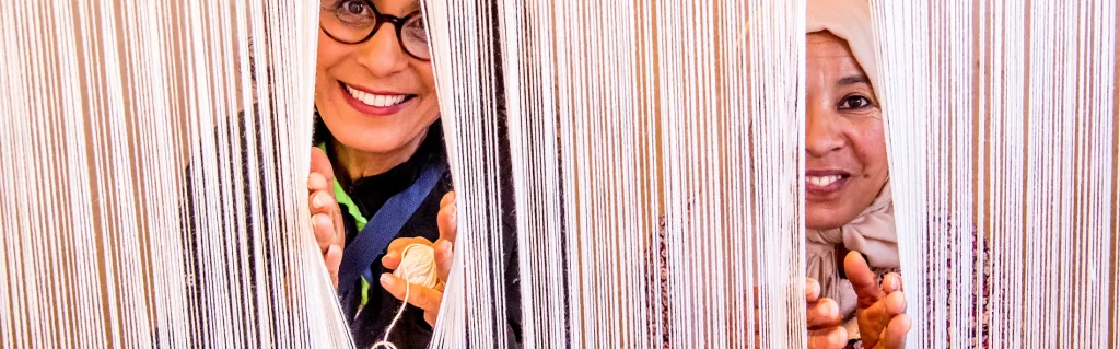 The image shows two women peering through a wall of white yarn or string, with their faces visible between the strands. * The woman on the left is wearing glasses and has her hands raised to her face as if she is trying to see something. * She is holding a ball of yarn in one hand. * Her mouth is open, suggesting that she may be smiling or laughing. * The woman on the right is also peering through the wall of yarn. * She appears to be looking at something with interest. * Her hands are not visible in this part of the image. The background of the image is a solid beige color, which helps to focus attention on the two women and their interaction with the yarn. Overall, the image suggests that the two women are engaged in some kind of creative activity or playfulness, possibly related to knitting or crochet.