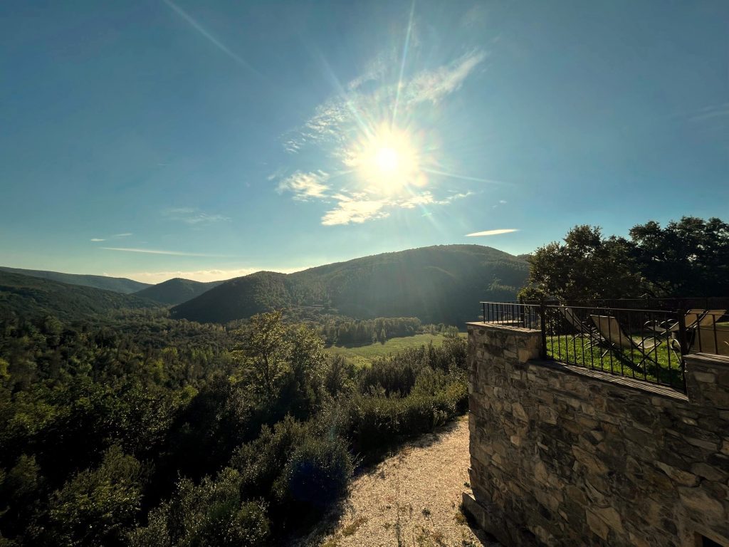 A panoramic view of rolling hills under a bright sun, with a stone wall and greenery in the foreground, creating a serene landscape.