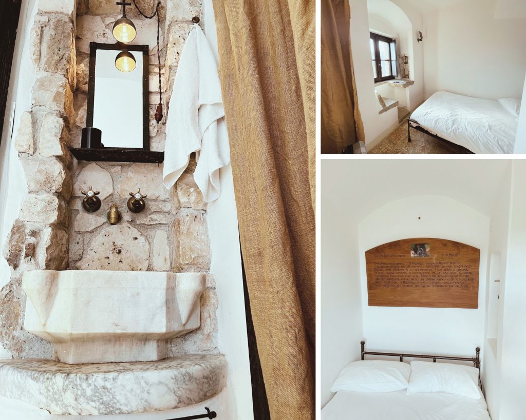 This photograph presents an interior view of a room, likely in a hotel or resort, showcasing various features. On the left, a stone wall with a built-in sink is prominently displayed, featuring two marble basins set into the wall, accompanied by three faucets and a mirror. The right side of the image reveals a single bed against a white wall, adorned with a headboard bearing an inscription on a wooden plaque. Above the bed, a small window allows natural light to enter the room. The overall aesthetic of the room is characterized by a harmonious blend of rustic stone walls and sleek, modern fixtures, creating a unique and inviting atmosphere.
