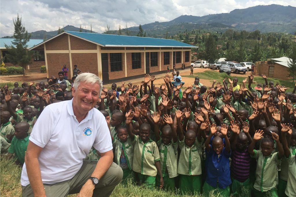 The image shows a man kneeling in front of a large group of children, with his hands raised in the air. The scene appears to be set in an outdoor location, possibly a school or community center. * A man is kneeling in front of a group of children: + The man has short white hair and is wearing a white polo shirt. + He is kneeling on one knee, with his other leg bent at the knee. + His hands are raised in the air, palms facing upwards. * The children are all wearing green uniforms: + They are standing behind the man, forming a semi-circle around him. + Each child has their arms raised in the air, mirroring the man's pose. + Their green uniforms appear to be identical, suggesting that they may be students at a single school or institution. Overall, the image suggests that the man is some kind of authority figure or leader, perhaps a teacher or principal, who is interacting with and inspiring the children. The fact that they are all raising their hands in unison adds to the sense of unity and cooperation among them.