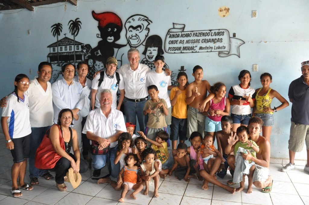 The image depicts a group of people, including children, posing for a photo in front of a wall adorned with graffiti-style artwork. In the foreground, two rows of individuals stand together, smiling at the camera. The first row consists of adults, while the second row comprises children and young teenagers, some barefoot and others wearing sandals or shoes. A few individuals wear white shirts, while others sport various colored tops and bottoms. One person in the back row is crouching down to be closer to a child. The background features a light blue wall with black graffiti-style artwork depicting palm trees, buildings, a cartoon drawing of a man's head, and text written in Portuguese. The overall atmosphere suggests a family or community event, possibly a gathering for children.
