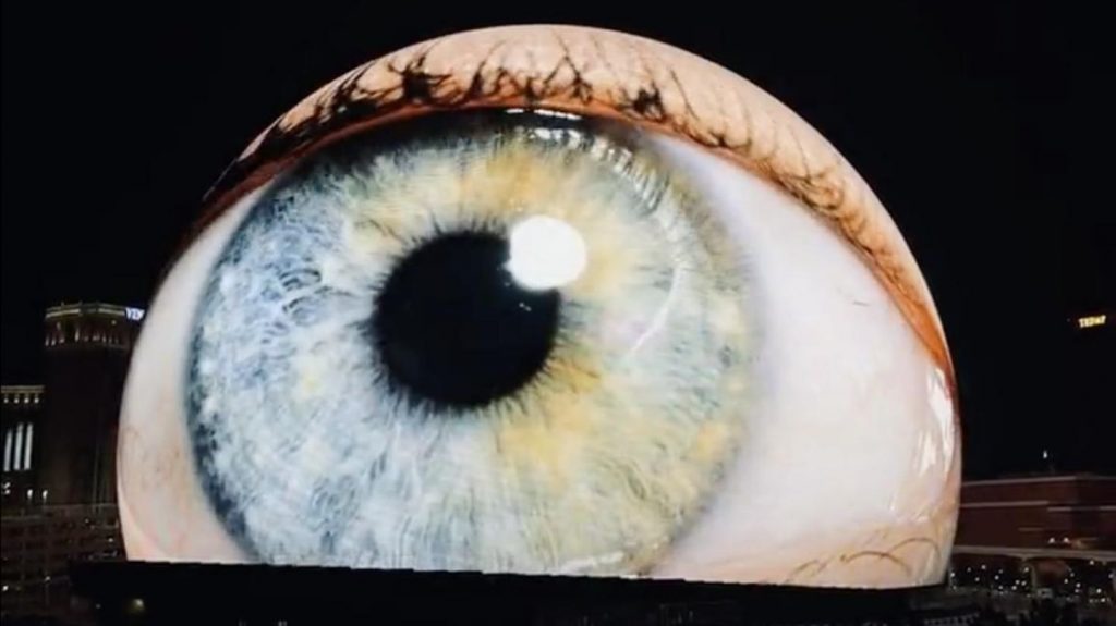A large, illuminated close-up of a human eye projected on a building at night, showcasing intricate details of the iris and eyelashes.