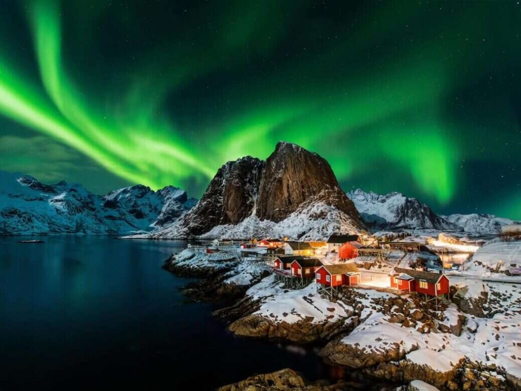 Alt tag: "A stunning night scene featuring the Northern Lights illuminating the sky over snow-covered mountains and a quaint village with red buildings by the water."