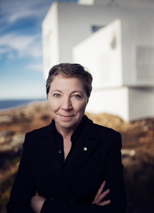 Confident woman in a black coat standing with arms crossed in front of a modern white building, with a blue sky and rocky surroundings in the background.