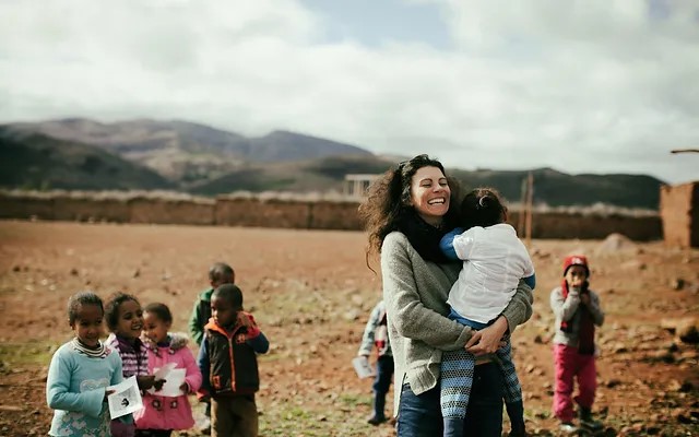 The image depicts a woman holding a child in her arms, surrounded by other children standing behind them. The scene is set against the backdrop of a mountain range. * A woman is holding a child in her arms: + The woman has long dark hair. + She is wearing a gray sweater and blue jeans. + Her facial expression is joyful, with a wide smile on her face. * There are several children standing behind them: + They appear to be of similar age to the child being held by the woman. + Some of them are looking at the camera, while others are gazing away. + They seem to be in a natural and relaxed environment. The overall atmosphere of the image suggests that it was taken during a happy moment or celebration, possibly in a rural or outdoor setting. The presence of the mountain range in the background adds a sense of grandeur and serenity to the scene.