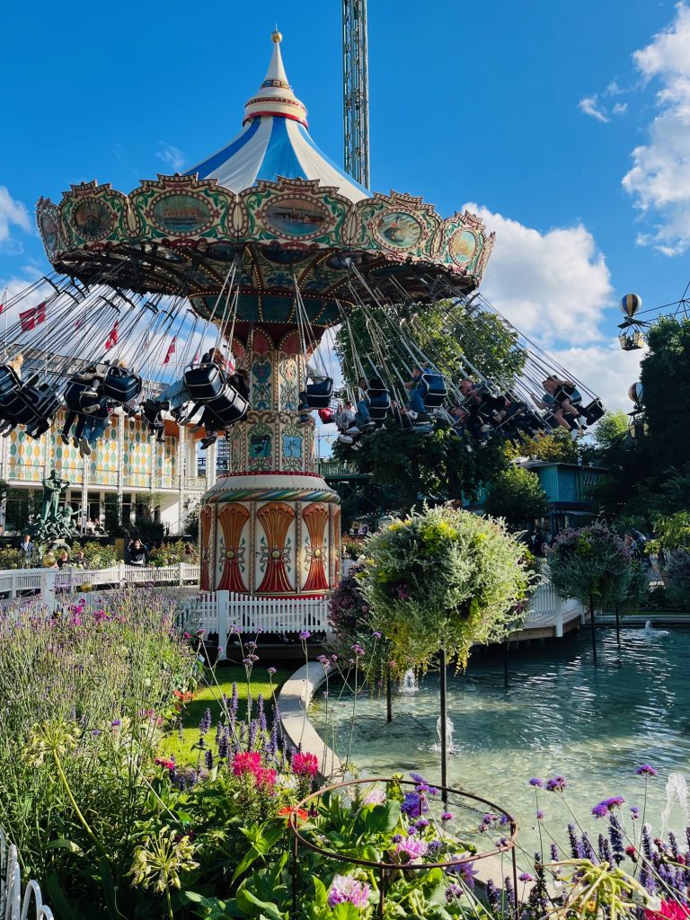 The image depicts a vibrant amusement park scene, featuring a prominent swing ride as its central attraction. In the foreground, a large body of water is surrounded by lush greenery, including trees and flowers. The swing ride itself boasts an ornate design, with a white and blue striped roof that adds to its visual appeal. Suspended from above are numerous swings, each adorned with black seats suspended by cables from the ride's structure. Beyond the main attraction, other rides can be seen in the background, while a bright blue sky dotted with clouds completes the idyllic atmosphere of this summer evening setting.