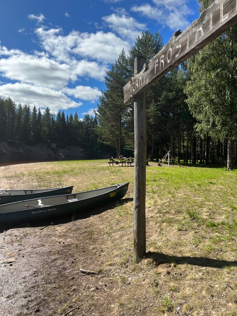 Alt tag: "A wooden sign reading 'Canoe Trail' stands near a sandy area with two canoes on the shore, surrounded by lush trees and under a partly cloudy sky."