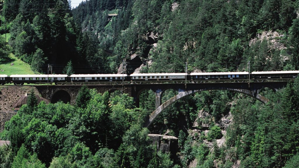 A scenic view of a train crossing a stone arch bridge amidst lush green trees and mountains.