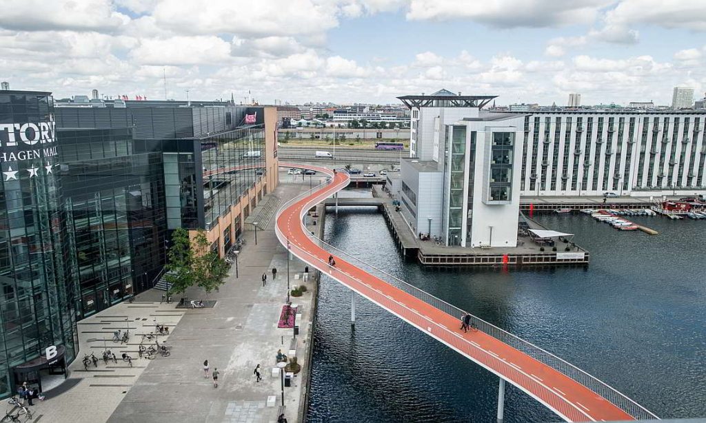 This image depicts a vibrant cityscape, with a prominent orange bike path running along the edge of a canal or river. In the foreground, a modern white building stands on the right side of the photo, featuring large windows and a flat roof. On the left side, a black building bears the words "HAGEN MALL" in white letters. Beyond these structures lies a bustling cityscape with numerous buildings and cars visible in the distance. The bike path, situated along the water's edge, is flanked by concrete walkways, where several people can be seen walking or standing. The canal itself is filled with dark blue water, adding to the urban landscape.