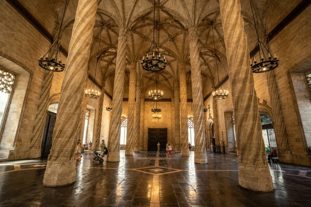 This image presents two photographs of an ornate, arched interior space. The top photograph showcases a spacious room with numerous stone pillars that support the vaulted ceiling. The walls are constructed from light-colored stone, while the floor is composed of dark brown tiles featuring white accents in a diamond pattern. The space is illuminated by natural light pouring in through large windows on the left side, and artificial lighting emanates from several chandeliers suspended from the ceiling. In the background, several individuals can be seen walking or standing. The bottom photograph offers a closer view of one of these stone pillars, which stands at least three times taller than an individual standing beside it. The pillar's base is wider at the bottom than at the top, where its edges are rounded and adorned with intricate carvings.