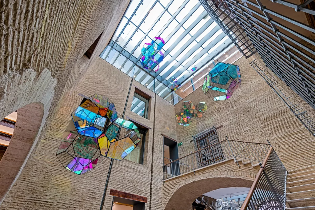 Interior view of a contemporary building featuring colorful, geometric glass sculptures suspended from the ceiling, with a textured stone wall and a skylight above.