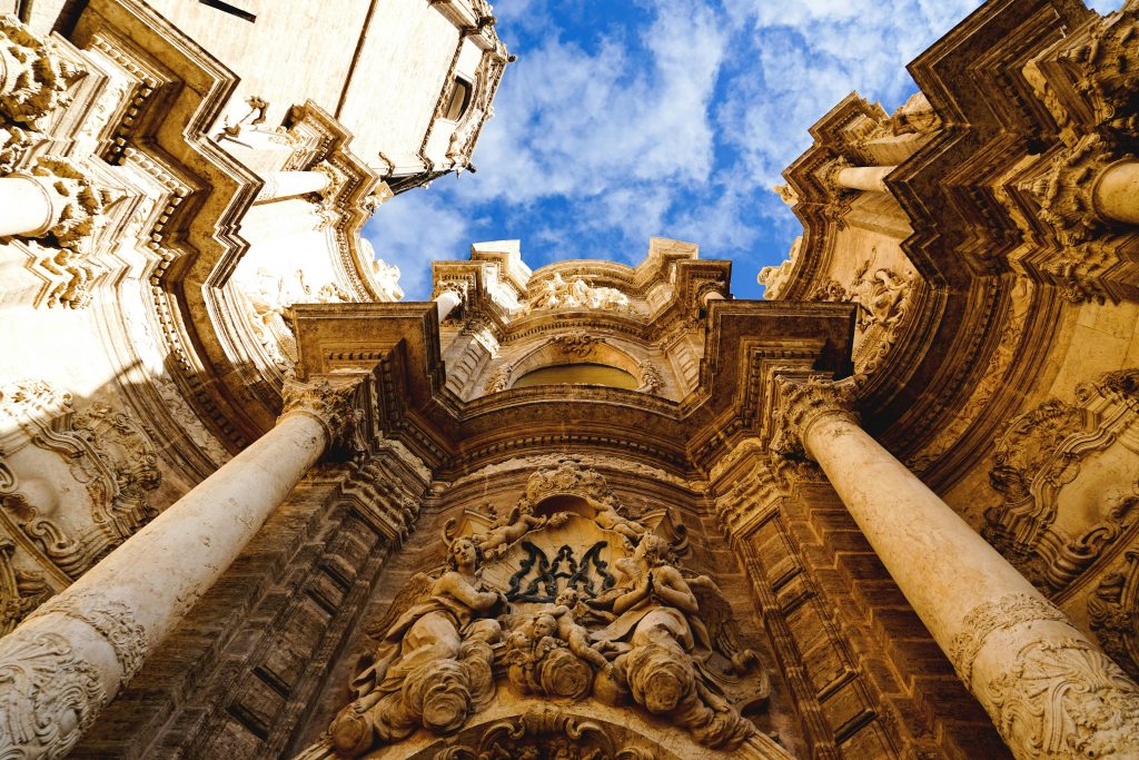 The image depicts the exterior of a church or cathedral, showcasing its ornate architecture. * The building's facade is adorned with intricate carvings and statues. + The carvings feature floral patterns, leaves, and other decorative elements. + The statues appear to be depictions of saints or biblical figures. * A large archway dominates the center of the image, flanked by two columns. + The archway is decorated with additional carvings and a central statue. + The columns are slender and feature intricate carvings at their bases. The image highlights the rich architectural details and ornate decorations that adorn this church or cathedral.