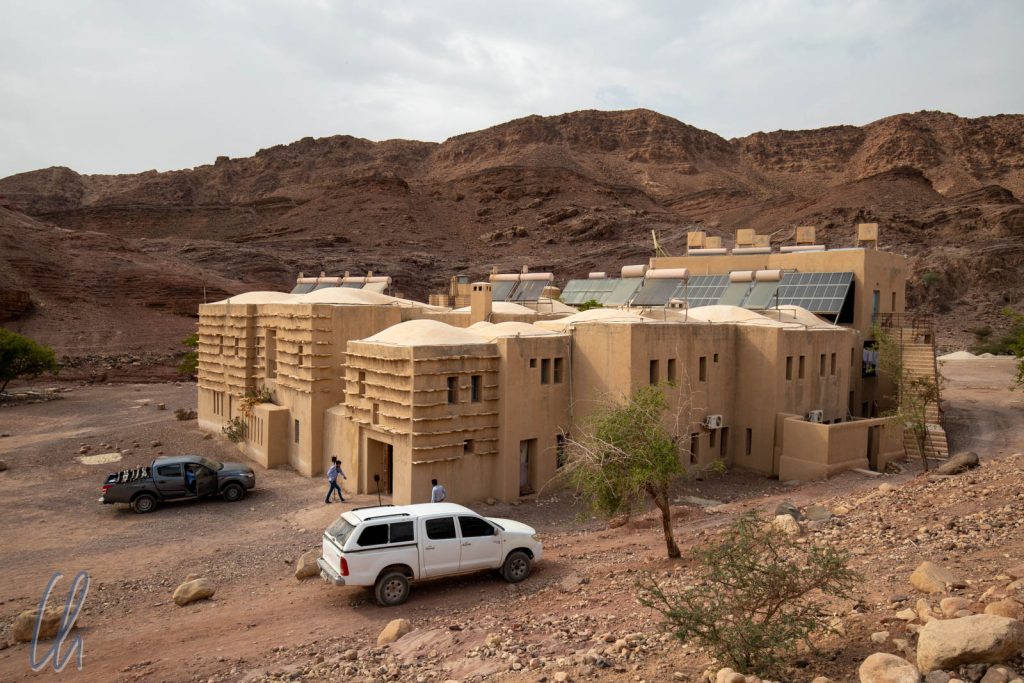 A modern adobe-style building surrounded by rocky terrain, with two vehicles parked in front and people walking nearby under a cloudy sky.
