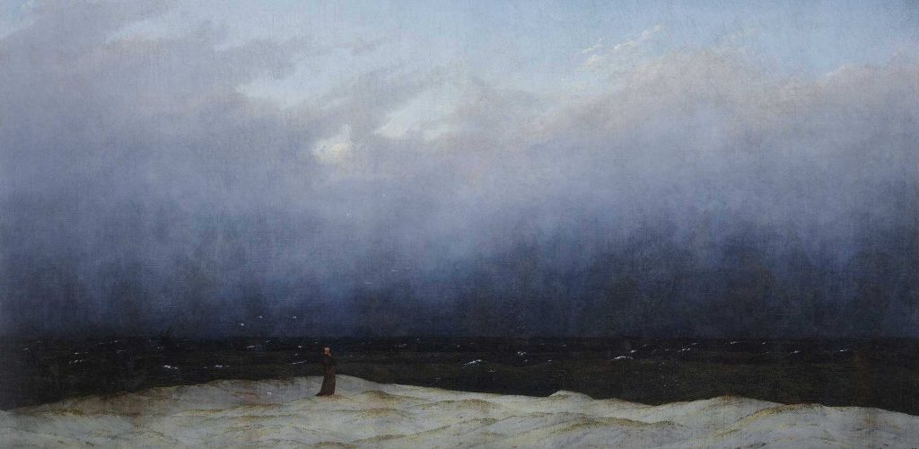 A solitary figure stands on a sandy shore, gazing out at a dark, turbulent sea under a cloudy sky, creating a mood of contemplation and isolation.