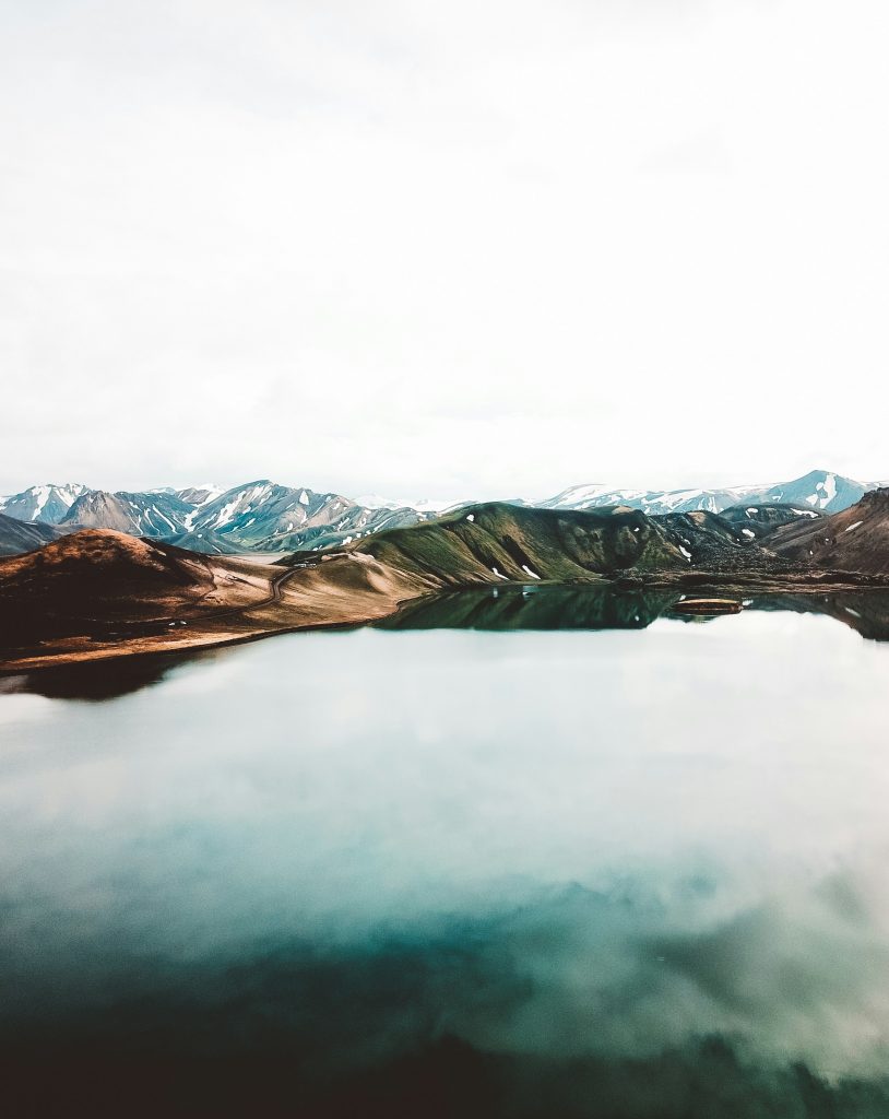 A serene landscape featuring a reflective lake surrounded by rugged mountains, with snow-capped peaks under a cloudy sky.