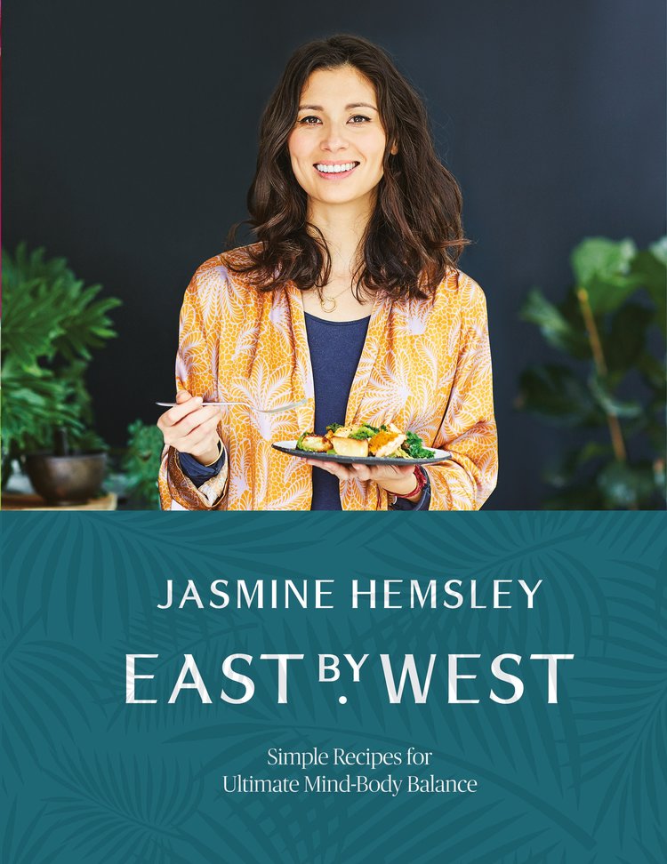Young woman smiling while holding a plate of food, wearing a colorful kimono-style robe, with green plants in the background; features the title and author name of the book "East by West" by Jasmine Hemsley.