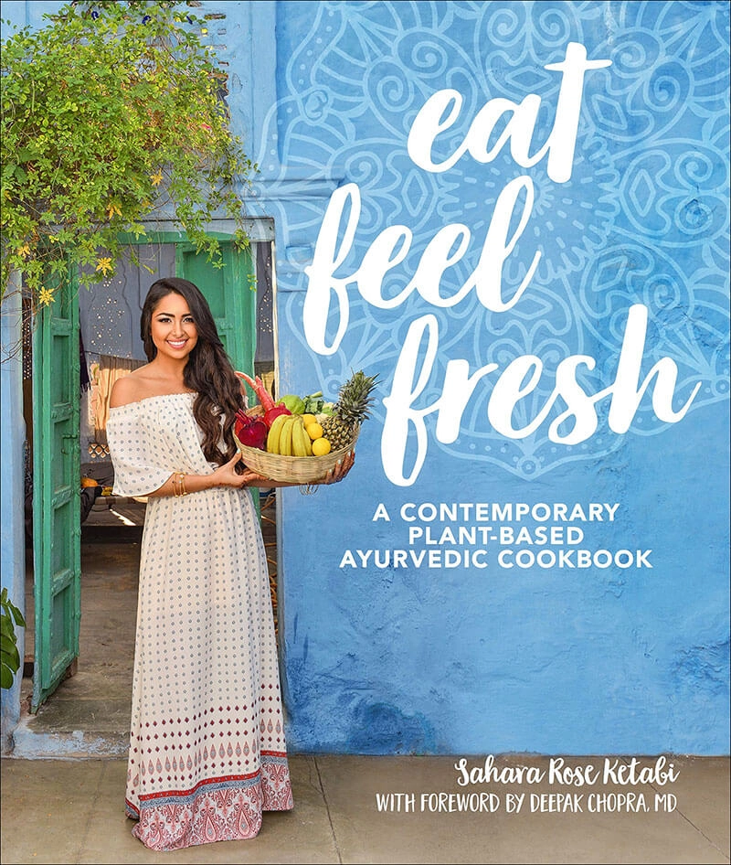 A smiling woman in a white off-shoulder dress holds a basket of fresh fruits in front of a colorful blue door, promoting the plant-based Ayurvedic cookbook titled "Eat Feel Fresh."