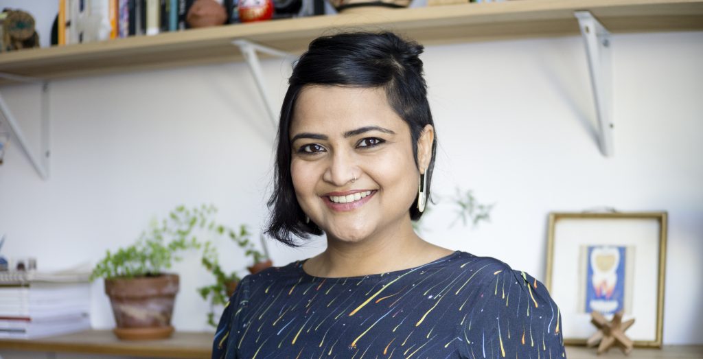 A smiling woman with short hair and a nose ring is standing in front of a bookshelf, wearing a patterned top and surrounded by green plants and decorative items.