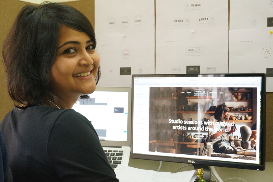A smiling woman sits at a desk, facing the camera, with two computer monitors displaying a website about studio sessions with artists. Design sketches are pinned on the wall behind her.