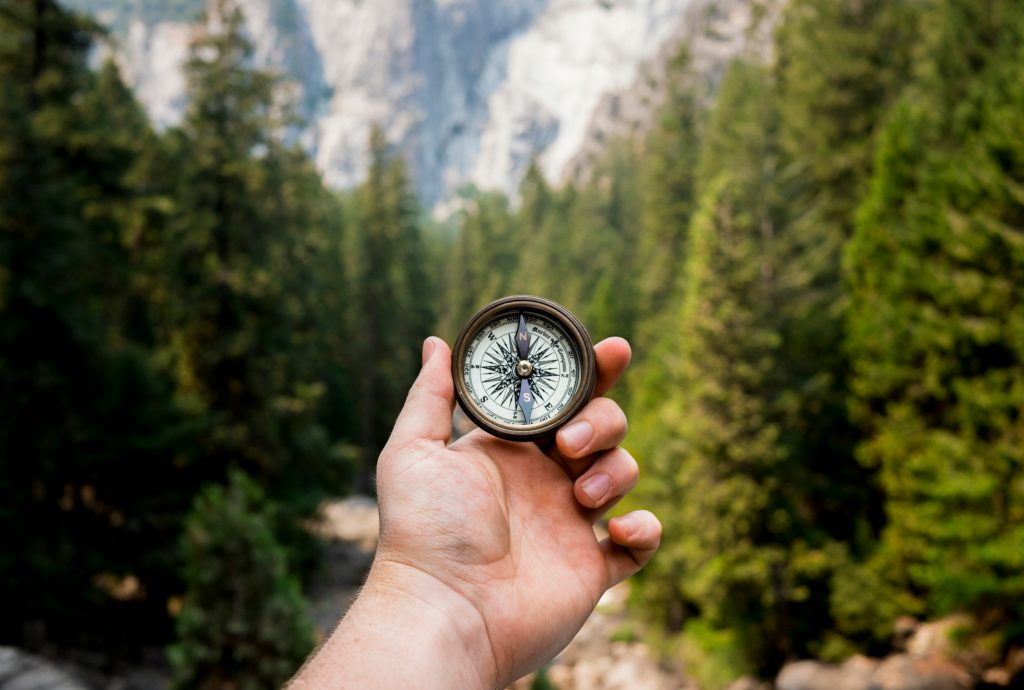 The image depicts a hand grasping a compass, set against the backdrop of a serene forest landscape. In the foreground, the hand is prominently displayed, with the palm facing upwards and the thumb on top of the compass. The thumb is positioned over the needle, which points towards one of the cardinal directions (north, south, east, or west). The rest of the fingers are wrapped around the compass to hold it securely in place. The background of the image features a dense forest with towering trees that stretch up towards the sky. The sunlight filters through the leaves, casting dappled shadows on the forest floor. In the distance, a rocky cliff face rises up, its rugged texture contrasting with the lush greenery below. Overall, the image presents a harmonious balance between the natural world and the tool used to navigate it.