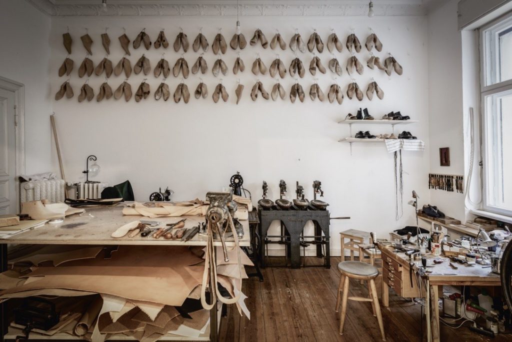 This photograph presents two images of a workshop, showcasing the interior of a room that appears to be dedicated to shoe-making or leatherwork. The top image features a wooden table with various tools and materials scattered across it, including pieces of wood, fabric, and other unidentifiable items. The wall above the workbench is adorned with numerous pairs of shoes hanging on hooks, adding a touch of elegance to the space. In the background, a black metal machine with three spindles is visible, alongside a small shelf holding additional tools. The bottom image provides a wider view of the room, revealing a large window along the back wall that allows natural light to pour in. The walls are painted white, and the floor appears to be made of dark wood or hardwood. A black metal machine with three spindles is situated next to the window, while several pairs of shoes hang on hooks above it.