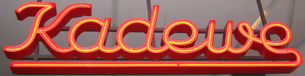 The image features a vibrant red neon sign with yellow accents, prominently displaying the word "Kade" in large cursive letters. In the foreground, the sign is illuminated by bright red lights that outline each letter, creating a striking visual effect. The word "Kade" is written in bold, flowing script, while the letters are connected to form a cohesive and dynamic design. At the bottom of the image, below the neon sign, lies a dark gray bar with a thin yellow line running along its length. The background of the image is not visible due to the close-up nature of the photograph, but it appears to be a wall or surface that provides context for the sign's placement and purpose. Overall, the image showcases a visually appealing and eye-catching neon sign that commands attention and draws the viewer in.