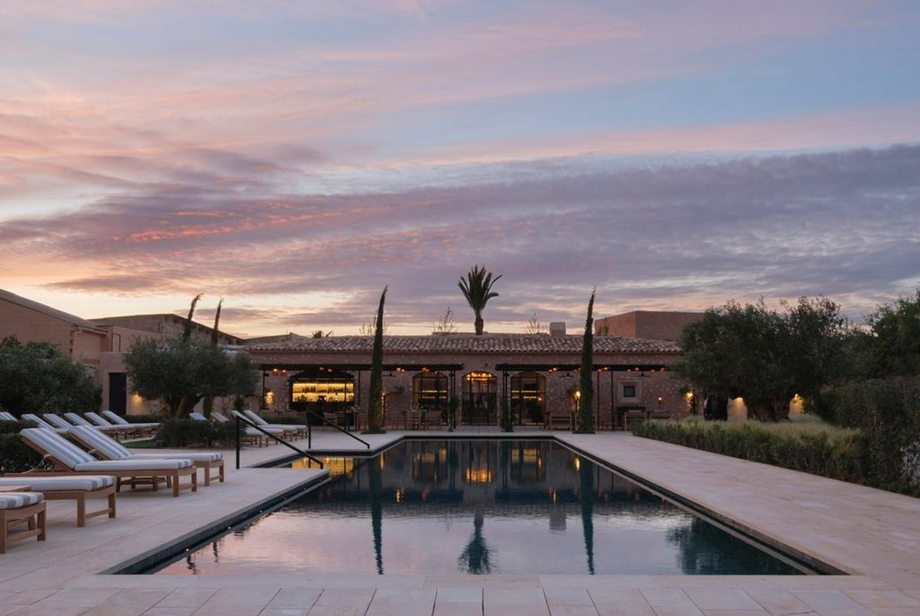 A luxurious outdoor pool area with lounge chairs, surrounded by lush greenery and a modern villa, set against a colorful sunset sky.