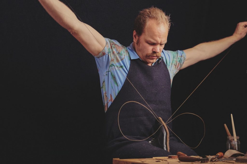 A focused artist in a floral shirt and apron, skillfully manipulating string while sitting at a workbench, surrounded by tools and materials.