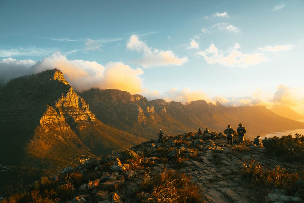 This photograph captures a breathtaking mountain range at sunset, with warm sunlight casting a golden glow on the rugged peaks. The rocky terrain is dotted with patches of greenery, while three individuals are visible in the distance, taking in the stunning view. The sky above is a brilliant blue, punctuated by wispy white clouds that add to the serene atmosphere. In the background, another mountain range stretches out, its contours softened by the fading light of day. The overall effect is one of natural beauty and tranquility, inviting the viewer to step into this idyllic scene.