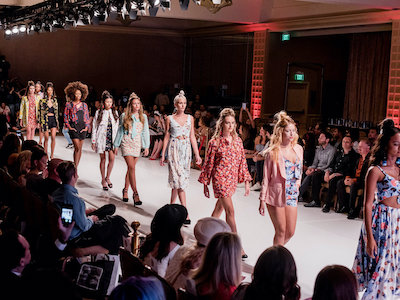 A group of models walking down a runway in front of an audience. The models are dressed in various outfits, including dresses, tops, and shorts, with some wearing heels and others flats. They are all facing forward, looking straight ahead as they walk along the catwalk. The background is filled with people sitting on chairs or standing up against walls, watching the show. The atmosphere suggests a fashion show or event, possibly a runway show for a clothing brand.