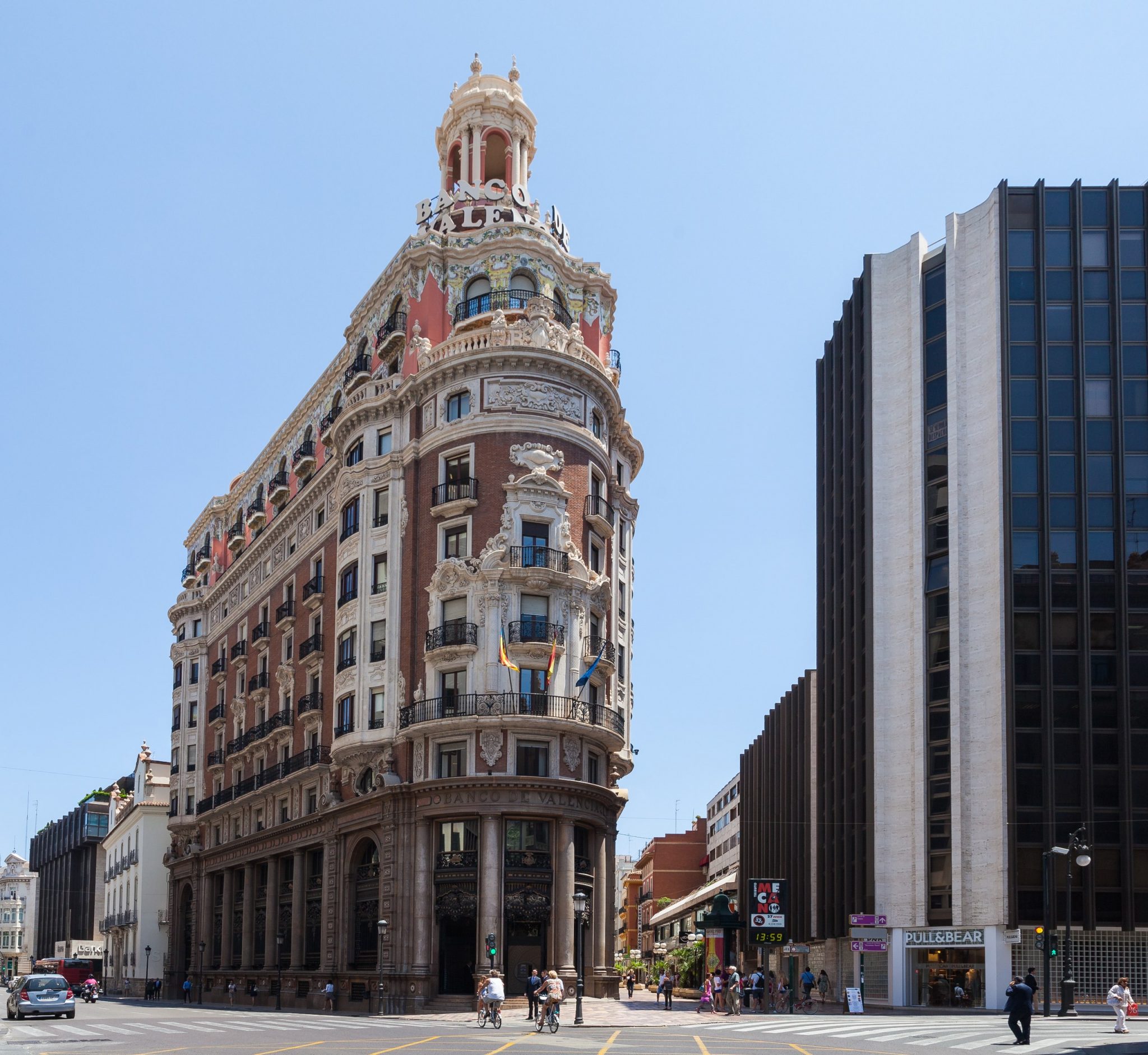 The image depicts a large, ornate building situated in the middle of an urban area, with a busy street and pedestrians visible in front of it. * **Building:** * The building is tall and rectangular in shape. * It has multiple stories, with at least five visible floors. * The exterior of the building features a mix of stone and brickwork. * There are several windows on each floor, some of which have balconies or terraces attached to them. * A large dome-shaped structure is located on top of the building, adding to its grandeur. * **Street:** * The street in front of the building is wide and straight, with a single lane for traffic in each direction. * There are several vehicles parked along the side of the road, including cars and buses. * Pedestrians can be seen walking across the street or standing on the sidewalk, some of whom appear to be waiting at bus stops or crossing points. * **Sky:** * The sky above the building is clear and blue, with no visible clouds or other obstructions. In summary, the image shows a large, ornate building located in an urban area, surrounded by a busy street and pedestrians.