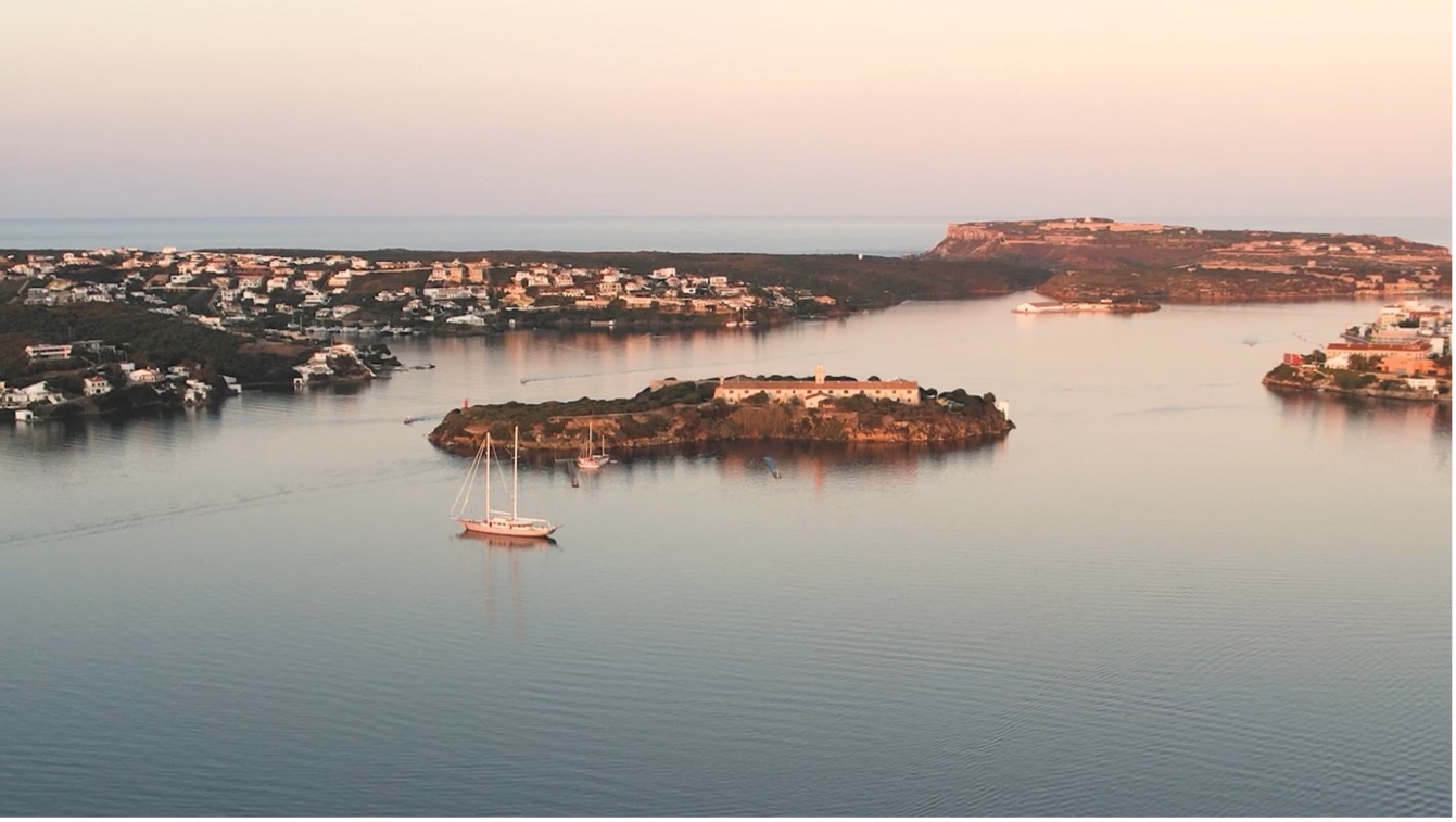 The image depicts a serene coastal scene, featuring small islands scattered across the water. In the foreground, two distinct islands are visible: one on the left with a cluster of buildings and another in the center-right, partially obscured by trees and foliage. The island on the right is entirely covered in vegetation, while the central island boasts a few structures along its shoreline. A sailboat navigates between these islands, adding to the tranquil atmosphere. The water surrounding the islands appears calm and peaceful, with gentle ripples that suggest a light breeze. Above, the sky transitions from yellowish-orange hues at the top left corner to a soft white towards the center of the image, gradually fading into a pale blue in the background. The overall ambiance exudes serenity and tranquility, inviting the viewer to immerse themselves in this idyllic coastal setting.