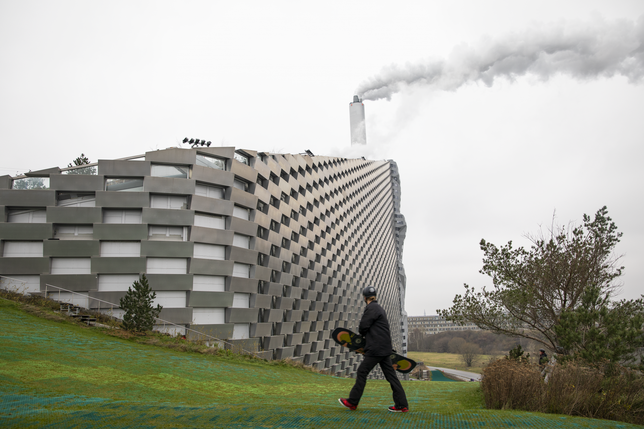 The image depicts a large, modern building with a unique design, featuring a series of interconnected cubes that form a curved wall. The building's exterior is composed of light-colored stone or concrete blocks, arranged in a staggered pattern to create a sense of depth and visual interest. In the foreground, a person wearing dark clothing and a helmet is walking towards the right side of the image, carrying a black skateboard with yellow wheels under their left arm. They appear to be heading towards the building, possibly to enter or exit it. The background of the image features a cloudy sky, suggesting that the photograph was taken on an overcast day. Overall, the scene conveys a sense of modernity and functionality, while also highlighting the beauty of architectural design.