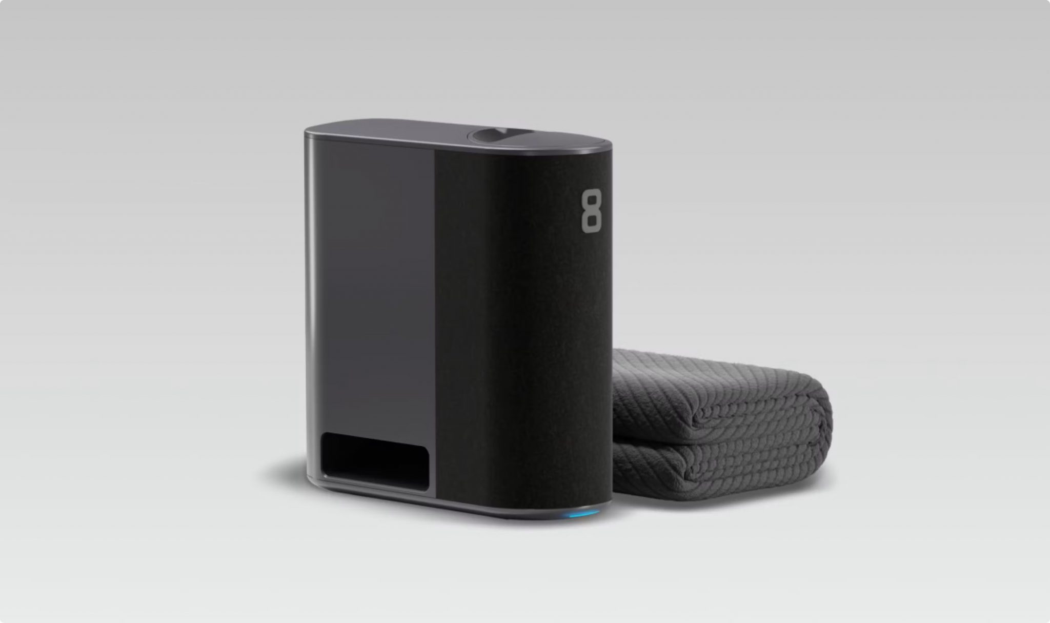 The image depicts a black, rectangular device with rounded edges, accompanied by two rolled-up blankets or towels to its right. * The device is positioned in the center of the image. + It has a flat top surface and a small circular indentation at the top. + A vertical strip on the front side features a white logo consisting of the number "8" followed by three smaller, indistinguishable shapes. + The bottom edge of the device glows blue. * To the right of the device are two rolled-up items that resemble blankets or towels. + They have a dark gray color with a textured pattern. + One item is slightly larger than the other and appears to be partially unrolled. The image suggests that the device may be some type of electronic appliance, possibly related to laundry or cleaning. The presence of the rolled-up items implies that they might be used in conjunction with the device for its intended purpose.