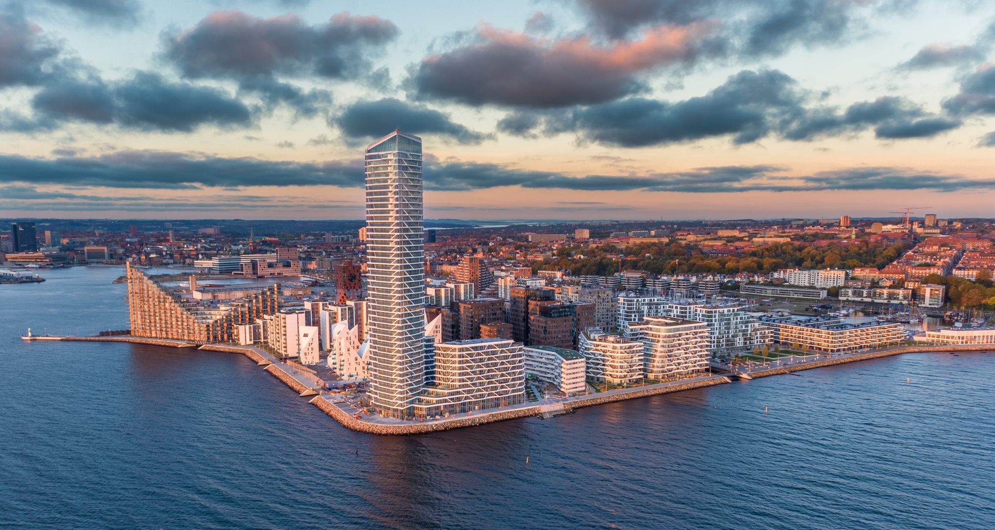 Aerial view of a modern waterfront city skyline featuring distinctive architectural buildings, including a tall, spiral tower, surrounded by water and green hills under a partly cloudy sky.