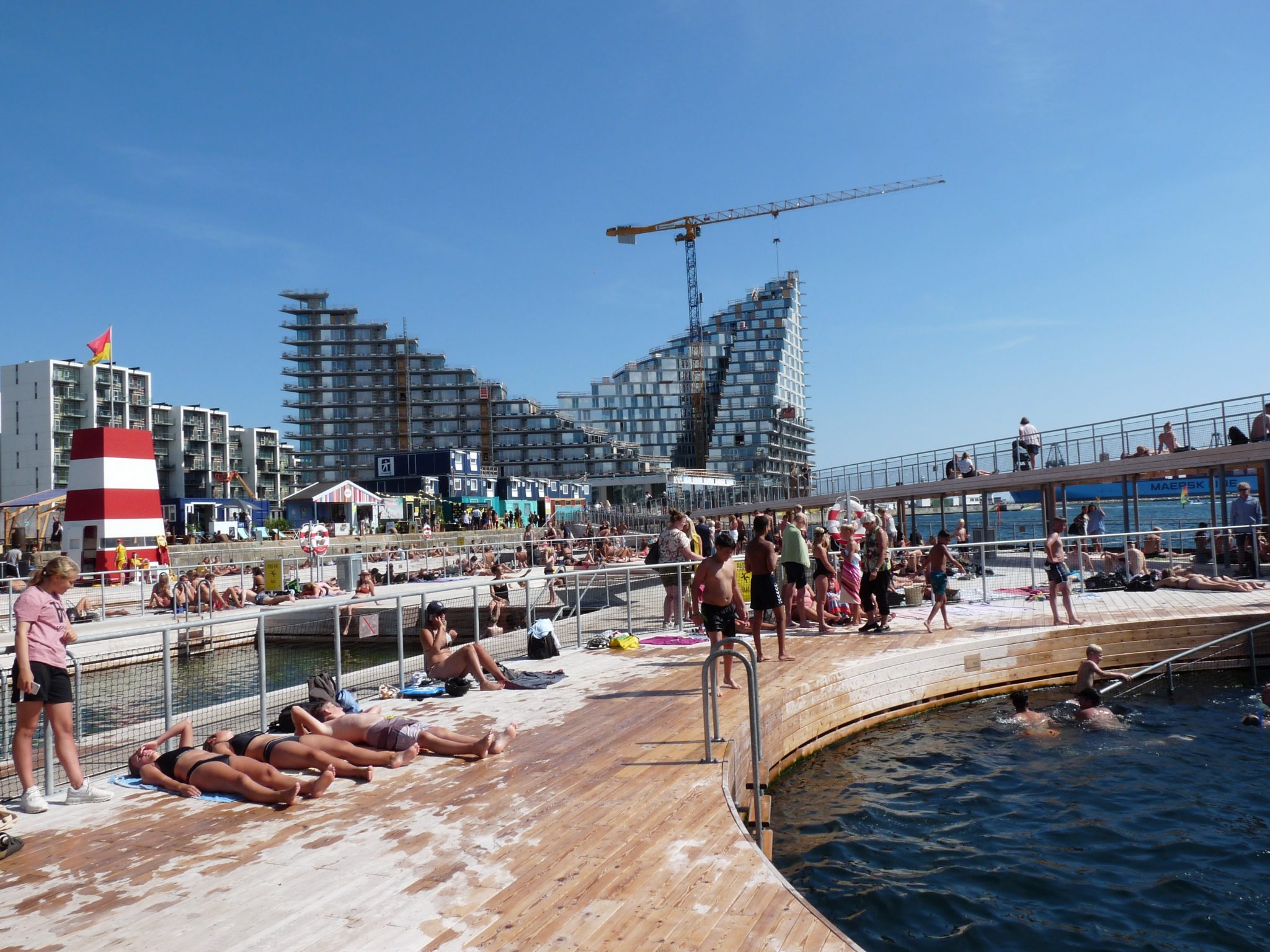The image depicts a bustling outdoor swimming pool area, with a large body of water in the foreground and a cityscape in the background. In the foreground, a group of people are lounging on the deck surrounding the pool, while others swim or float in the water. The pool appears to be a public facility, given its central location and the number of patrons present. Beyond the pool area, several large buildings are visible, including what appears to be a hotel or apartment complex under construction. A red-and-white striped object stands out in the distance on the left side of the image, adding a pop of color to the scene. The overall atmosphere suggests a warm summer day, with people enjoying the outdoors and soaking up the sun. The presence of a swimming pool and nearby buildings implies that this is likely an urban or suburban setting.