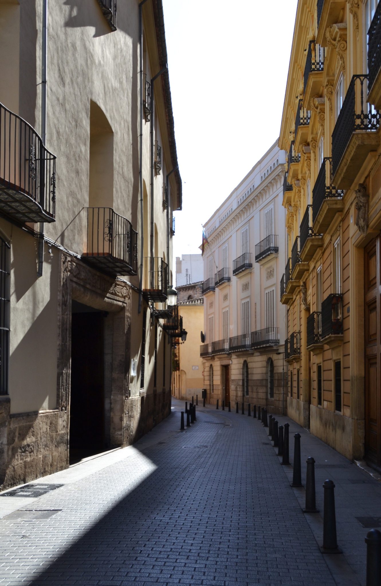 This image captures a serene, sun-dappled narrow street in an old city, lined with tall buildings that evoke a sense of history and tradition. The street's cobblestone or brick paving is interrupted by short bollards, while the buildings on either side feature ornate ironwork balconies and window frames. The façades are painted in warm shades of beige and yellow, with some displaying intricate stonework details. The overall ambiance suggests a quiet, tranquil setting, possibly during early morning or late evening hours when the sun casts long shadows along the street. The image exudes a sense of peace and serenity, inviting the viewer to step into this charming urban scene.