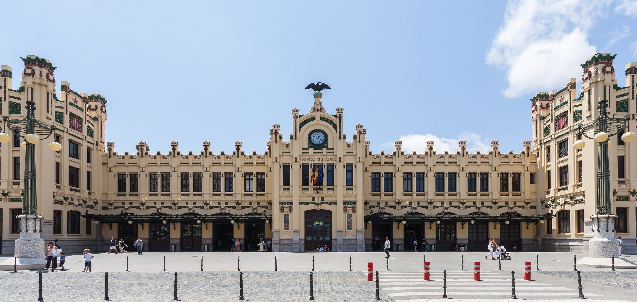 Historic train station facade featuring ornate architectural details, a central clock, and decorative elements, set against a clear blue sky with people walking in the plaza.