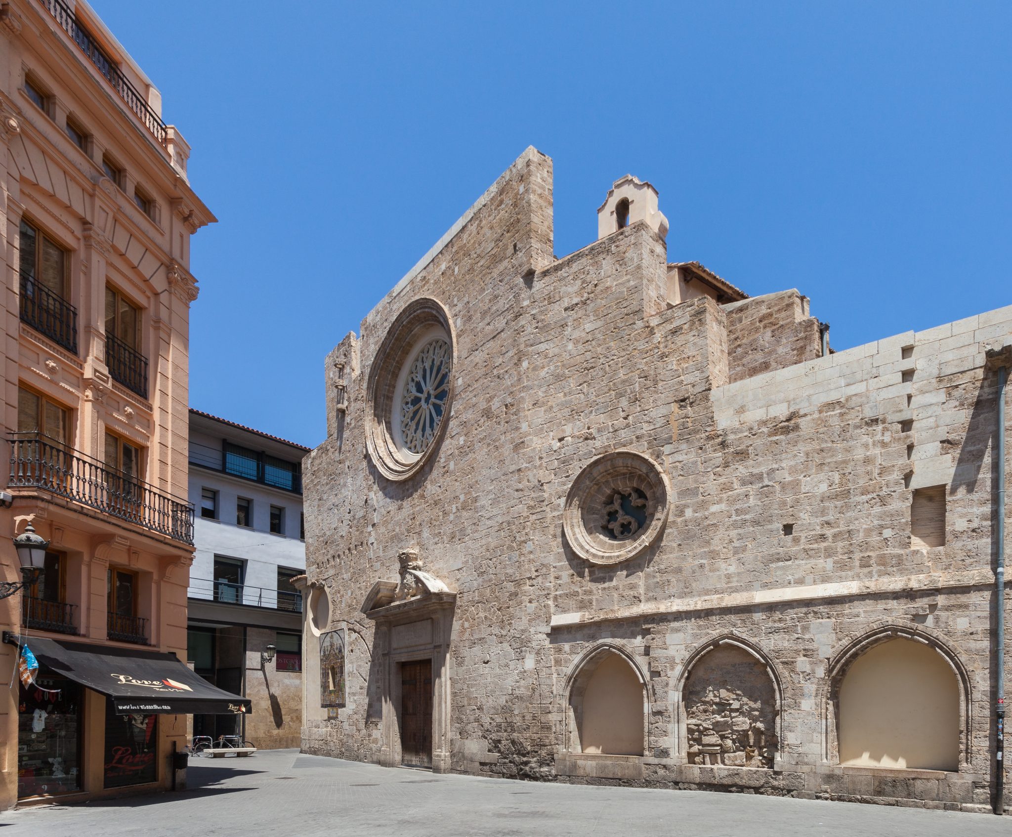 The image depicts the exterior of an old stone church, situated in a quiet street between two buildings. The building itself is constructed from light-coloured stones, featuring multiple arched windows on the lower level and a large round window with intricate stonework above it. The stone façade has several small openings near the top, possibly for ventilation or as decorative elements. In front of the church lies a narrow street, flanked by tall buildings that are at least two stories high. The overall appearance of the scene suggests a peaceful and serene atmosphere, evoking the sense of a quiet morning in an old European town.