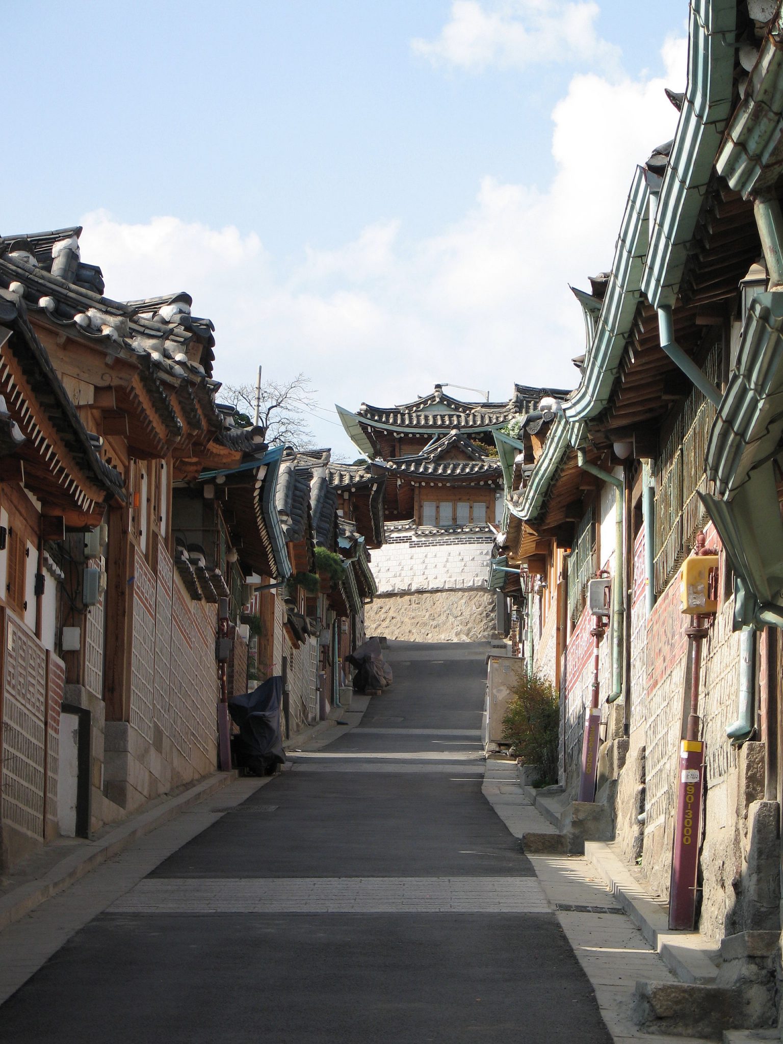 Alt tag: "Narrow traditional street lined with Korean hanok houses, featuring curved roofs, leading up to a distant building under a blue sky."