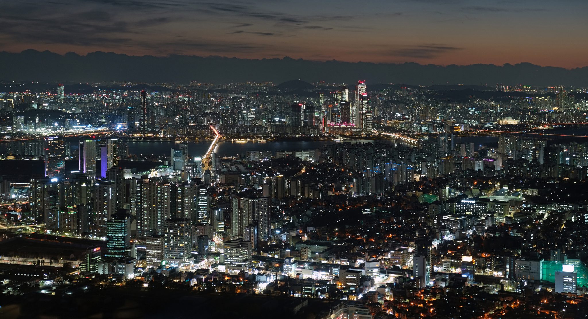 This nighttime cityscape showcases a sprawling metropolis, with numerous buildings of varying heights, some reaching into the sky, illuminated by an array of lights that cast a warm glow. In the foreground, a prominent structure stands out, while in the background, a mountain range stretches across the horizon, partially obscured by clouds. The sky is painted with hues of orange and purple, as if reflecting the vibrant colors of the city below.