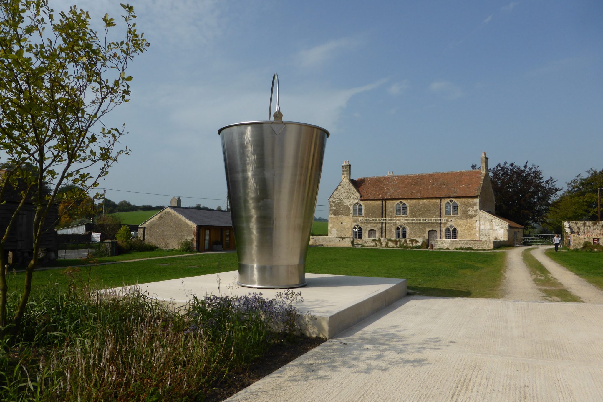 A large metallic bucket sculpture stands on a concrete base, surrounded by greenery and facing a stone building with a red-tiled roof, under a clear blue sky.
