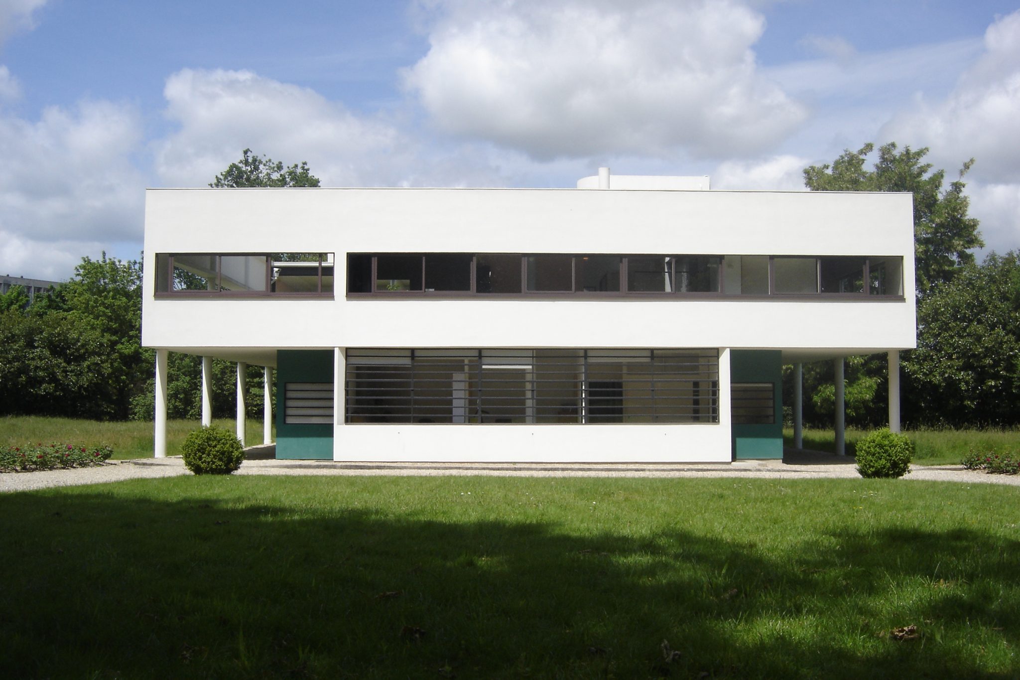 Alt tag: Modernist white building with large windows, supported by slender columns, set in a green landscape under a partly cloudy sky.