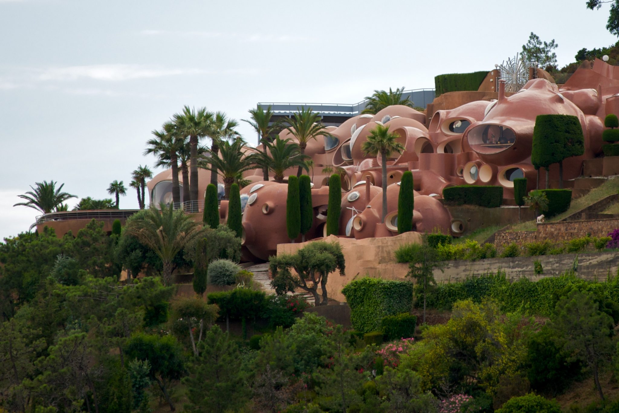 Modern, organic architecture with rounded, pink structures nestled among lush greenery and palm trees on a hillside.