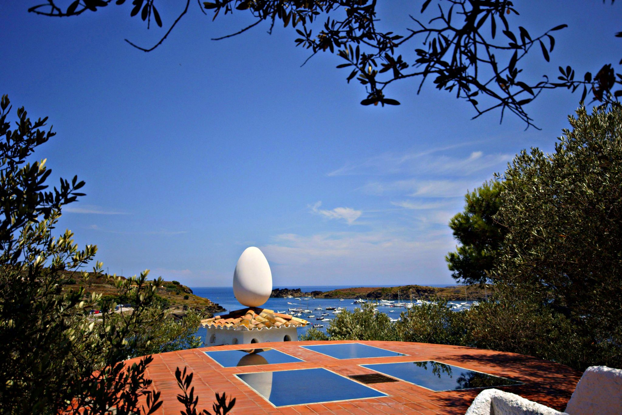 This photograph captures the stunning view of the Mediterranean Sea from a serene patio area. The foreground features a vibrant orange-tiled patio with three mirrored square pools, surrounded by lush greenery. In the distance, a majestic white structure stands on a rocky outcrop, its egg-like shape evoking an artistic flair reminiscent of Salvador Dali's surrealist style. The horizon stretches across the calm blue sea to meet the light blue sky dotted with wispy clouds. The overall ambiance exudes tranquility and serenity, inviting the viewer to bask in the beauty of this picturesque scene.