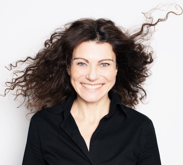 The image features a woman with long, curly brown hair that appears to be blowing in the wind, although it is likely manipulated using photo editing software. Her dark brown locks cascade down her back, framing her face. In the foreground, the woman wears a black collared shirt with a V-neckline and buttons at the top of the collar, adding a touch of sophistication to her overall appearance. The background of the image is a clean white, providing a neutral backdrop that allows the viewer's attention to focus on the subject. Overall, the composition of the image creates a sense of elegance and poise, with the woman's hairstyle and attire contributing to a polished look.