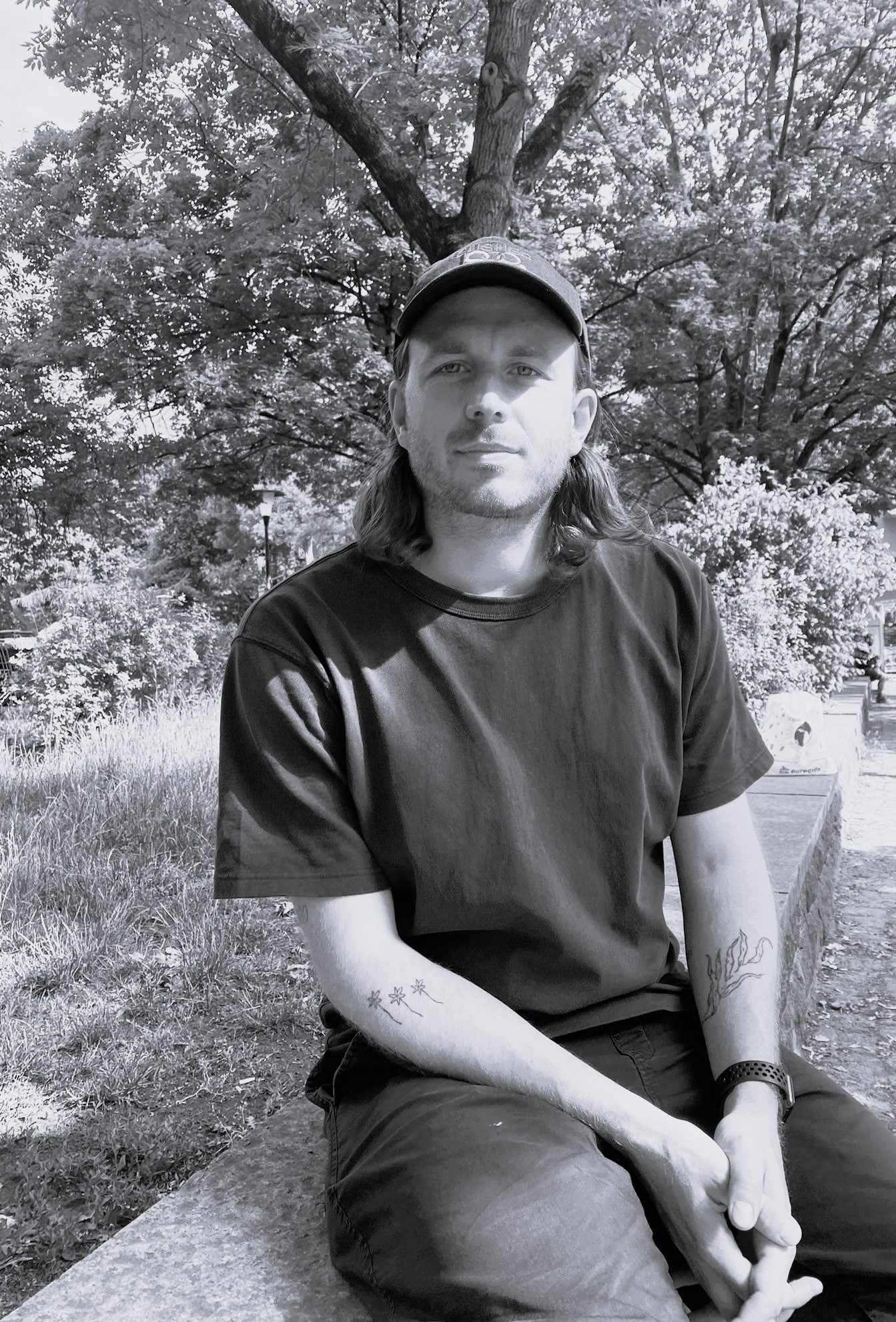 The image is a black-and-white photograph of a man sitting on a bench in front of trees. * The man is wearing: + A dark-colored t-shirt + A baseball cap + Dark jeans * He has tattoos on his arms, including one that appears to be a leaf or flower design on his right forearm and another that resembles a word or phrase on his left bicep. * The background of the image shows: + Trees with green leaves + Grass The overall atmosphere of the image suggests a casual, relaxed setting, possibly in a park or outdoor recreational area.