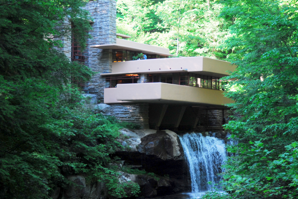 The image depicts the exterior of Fallingwater, a renowned residence designed by architect Frank Lloyd Wright. **Key Features:** * The house is situated on a rocky outcrop overlooking Bear Run Creek. * A waterfall cascades underneath the cantilevered deck at the rear of the house. * The building's design seamlessly integrates with its natural surroundings, featuring: + Large windows and sliding glass doors that connect the interior to the outdoors. + Exposed beams and wooden accents throughout the structure. **Overall:** The image showcases Fallingwater as a masterpiece of organic architecture, exemplifying Wright's philosophy of harmonizing buildings with their environment.