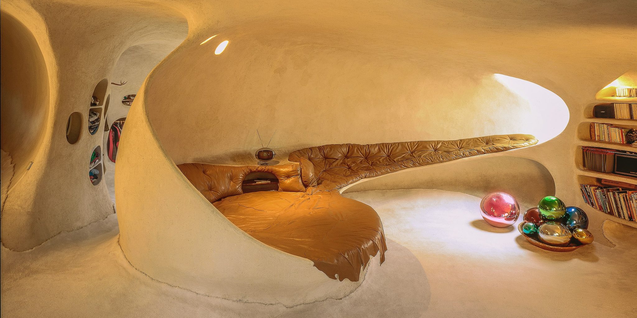 The image presents a unique, organic-shaped room with curved walls and ceiling, featuring a circular bed nestled in the center. * The room's interior is characterized by its wavy, irregular shape, creating a sense of fluidity and dynamism. * The walls are painted white, providing a clean and neutral backdrop for the surrounding decor. * A small window on the left side allows natural light to enter the room, adding warmth and depth to the space. * In the center of the room, a circular bed is situated, surrounded by a soft, golden-colored headboard that adds a touch of elegance to the overall design. * The bed itself appears to be made of a plush material, inviting relaxation and comfort. * A small nightstand or dresser sits beside the bed, providing storage for personal items and adding to the room's functionality. * On the right side of the room, a bookshelf stands tall, filled with books that add a sense of coziness and intellectual curiosity to the space. * The bookshelf is designed with rounded edges, blending seamlessly into the wavy shape of the room. * A small lamp on top of the bookshelf provides warm lighting, creating a cozy reading nook. * In front of the bed, a circular rug adds warmth and texture to the space, complementing the soft colors used throughout the room. * The rug's design features a subtle pattern, adding visual interest without overwhelming the senses. * A small table or stool sits in front of the rug, providing a convenient surface for placing items such as books, glasses, or a phone. In summary, this image showcases a unique and inviting room with an organic shape, featuring a circular bed, bookshelf, and other decorative elements that create a cozy and functional space.
