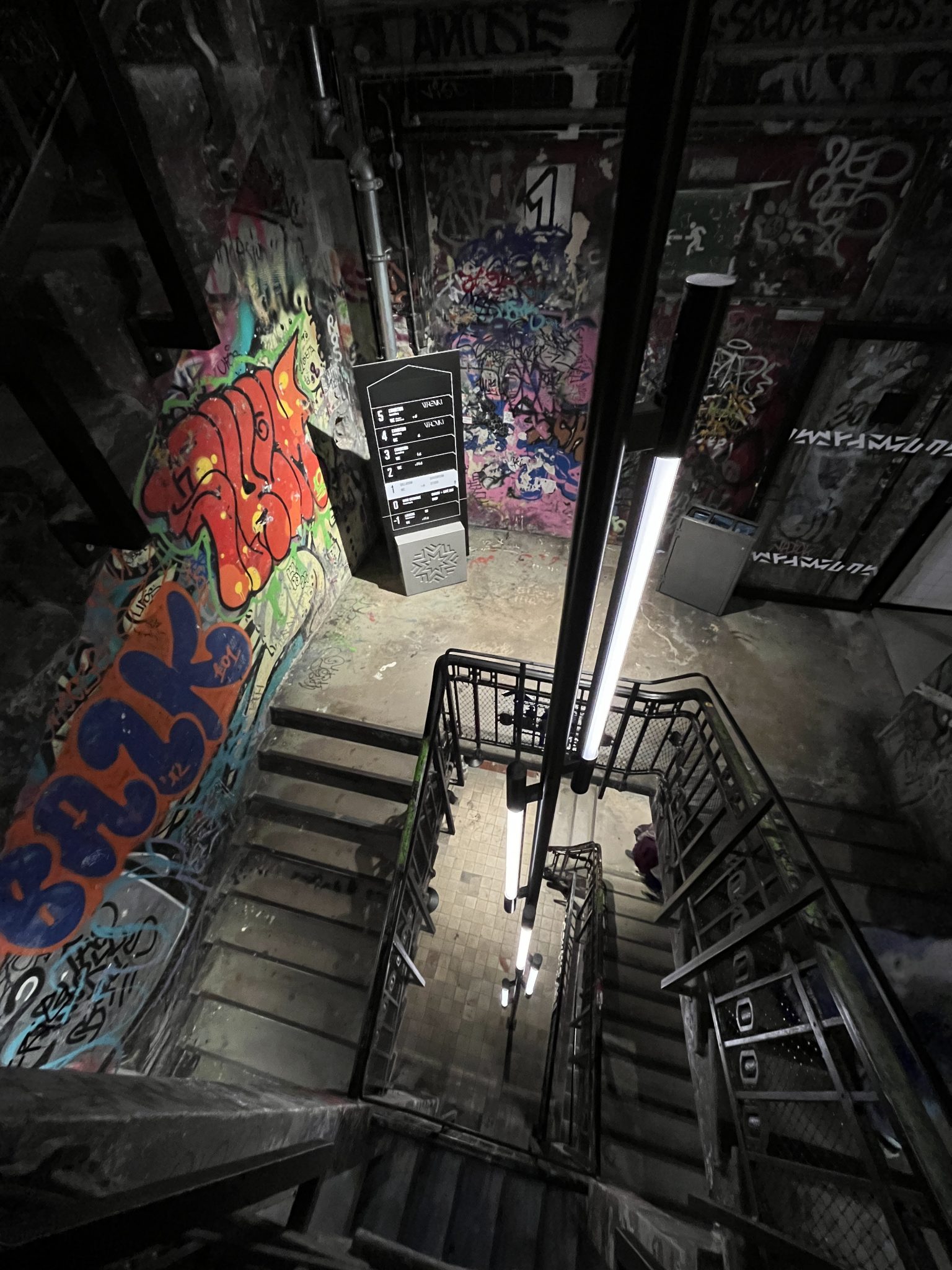 The image depicts a dimly lit, narrow staircase within a building that appears to be in disrepair. The walls are covered in vibrant graffiti and murals, adding a pop of color to the otherwise drab atmosphere. **Key Features:** * **Staircase:** Narrow and dimly lit, with metal railings on both sides * **Walls:** Covered in colorful graffiti and murals, creating a lively contrast to the surrounding environment * **Flooring:** Appears to be made of concrete or stone, with visible signs of wear and tear * **Lighting:** Dim, with only a few lights illuminating the area, casting long shadows across the walls **Atmosphere:** * **Abandoned:** The building appears to be in disrepair, with broken windows and peeling paint * **Artistic:** The vibrant graffiti and murals suggest that the space has been used as an art studio or gallery at some point * **Industrial:** The metal railings and concrete flooring evoke a sense of industrial heritage **Overall:** The image presents a unique blend of neglect and creativity, with the colorful artwork standing out against the drab, worn surroundings. The dim lighting adds to the atmosphere of abandonment, while the metal railings and concrete flooring hint at the building's industrial past.